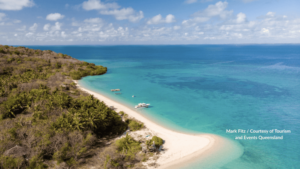 Haggerstone Island in Far North Queensland with curved white beach and turquoise reef flats with a boat along the palm shoreline