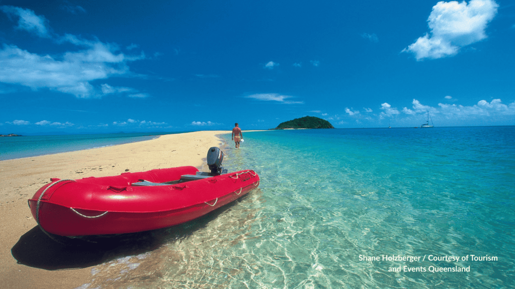 Red dinghy on Langford Island sand spit in the Whitsundays Queensland with a visitor walking the white sand toward the island cay and a yacht offshore